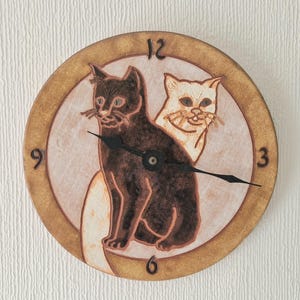 May include: Circular ceramic wall clock with two cat illustrations. The clock face has a light brown background, a darker brown border, and black clock hands. The cats are brown and cream-coloured.