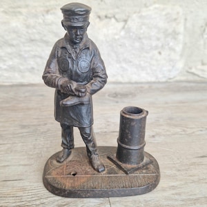 Puede incluir: Un antiguo sujetador de cerillas de hierro fundido que representa a un hombre uniformado sosteniendo un zapato. La figura de metal oscuro se encuentra sobre una base rectangular con un portacerillas cilíndrico. La pieza tiene un aspecto rústico y envejecido.