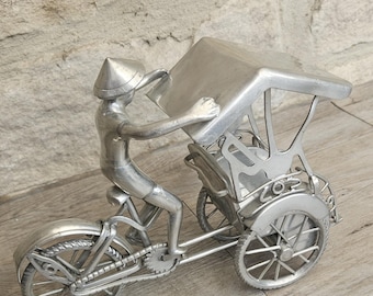 May include: A silver metal sculpture of a cyclo, a three-wheeled bicycle taxi. The cyclo features a driver wearing a conical hat, pedalling and holding the passenger compartment's roof. The sculpture is detailed with spokes, gears, and a seat.