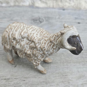 May include: A ceramic sheep figurine with a textured, off-white woolly body and a dark brown face. The sheep has curved horns and is posed in a standing position. The figurine is set against a light gray wooden surface.
