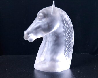 Puede incluir: Una escultura de cristal transparente con forma de cabeza de caballo. La cabeza está detallada, con una melena fluida y una oreja puntiaguda. La escultura se encuentra sobre un fondo oscuro.