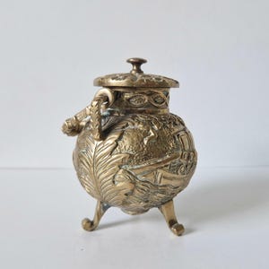 Puede incluir: Una pequeña olla de latón antigua con tapa y tres patas curvas. La olla presenta intrincados diseños en relieve, incluyendo hojas y un edificio. El asa también está decorada con un diseño de hojas.