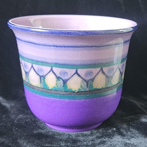 May include: A hand-painted ceramic cup with a flared rim. The cup features a gradient of colors, transitioning from lavender to purple. A decorative band with teal, gold, and blue accents encircles the middle. The cup is a unique piece of art.