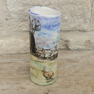 May include: A cylindrical ceramic vase with a hand-painted landscape scene. The design features a windmill, a small house, and a running animal against a backdrop of a blue and white sky. The vase is approximately 6 inches tall.
