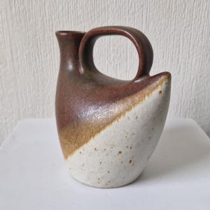 Puede incluir: Una pequeña jarra de cerámica marrón y blanca con una forma redondeada única y un asa. La jarra tiene un esmalte moteado.