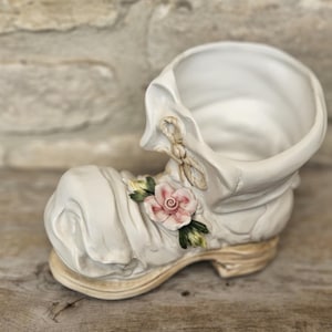 May include: A white ceramic boot-shaped planter with a pink rose and green leaves. The boot has a textured surface and a beige sole. The planter is designed to hold small plants or flowers.