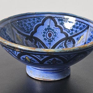 May include: A blue ceramic bowl with detailed floral patterns in white, black, and yellow. The bowl has a pedestal base and a slightly worn rim. The interior design features a central medallion and symmetrical motifs.