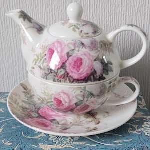 Puede incluir: Un juego de té de porcelana blanca con tetera, taza y platillo. El juego está decorado con motivos florales de rosas rosas y follaje verde. La tetera tiene tapa y asa, y la taza se encuentra dentro de la tetera.