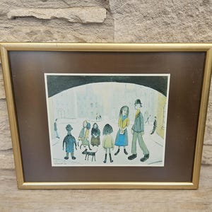May include: A framed print featuring a stylized illustration of several figures and a small dog standing in a city square. The artwork uses muted tones of blue, yellow, and green, and is enclosed in a thin gold-colored metal frame with a brown mat.