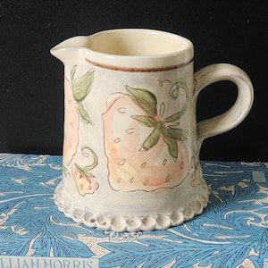 May include: A ceramic pitcher with a cream-colored body and a handle. The pitcher is decorated with hand-painted strawberries in shades of pink, green, and brown. The base has a scalloped edge.