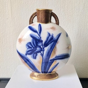 May include: A round, white ceramic vase with blue and gold floral designs. The vase has two handles and a gold base.