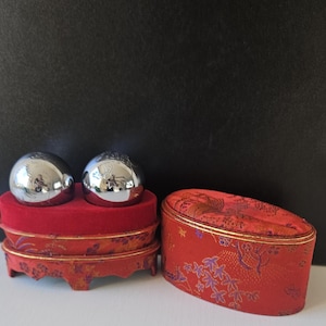 May include: Two polished silver spheres sit on a red velvet platform with a decorative, tiered base. Beside them is a red, oval-shaped box with a floral pattern and gold trim. The background is a dark, solid colour.