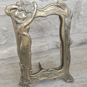 May include: A rectangular brass picture frame featuring a sculpted figure of a person with flowing hair on the left side. The frame is decorated with embossed floral and leaf motifs along the edges and includes a metal stand on the back for support.