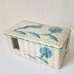 以下が含まれることがあります： 蓋付きの四角いセラミックボックスで、薄いベージュの背景に青と緑の花柄が描かれています。ボックスの側面には小さな窓があります。
