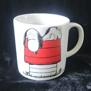 May include: White ceramic mug with a cartoon illustration of Snoopy asleep on his red doghouse. The mug has a curved handle. The design includes black and white details and is set against a dark background.