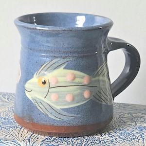 May include: A blue ceramic mug with a hand-painted fish design. The fish is yellow with pink spots and black details. The mug has a brown base and a dark blue handle. The mug is suitable for hot or cold drinks and measures approximately 10 cm in height.