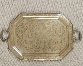 Puede incluir: Una bandeja octogonal plateada con diseños grabados y dos asas curvas. La bandeja presenta patrones detallados de figuras y motivos florales, adecuada para servir o exhibir.