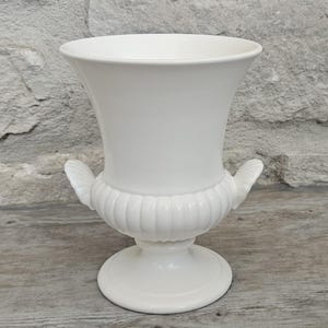 May include: A white ceramic urn-style vase featuring a wide, flared rim, a rounded body with vertical fluted detailing, and two small, shell-shaped handles on either side. The vessel stands on a circular pedestal base with a smooth finish.