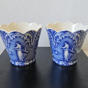 Puede incluir: Dos macetas de cerámica azul y blanca con borde festoneado. Cada maceta presenta un diseño clásico con una mujer, pájaros y motivos florales. Las macetas son probablemente vintage o antiguas.