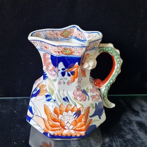 May include: A vintage, hand-painted ceramic pitcher with a unique, multi-faceted shape. The pitcher features a dragon-shaped handle with red and green accents. The body is decorated with orange, blue, and pink floral patterns, and the rim has a scalloped edge.