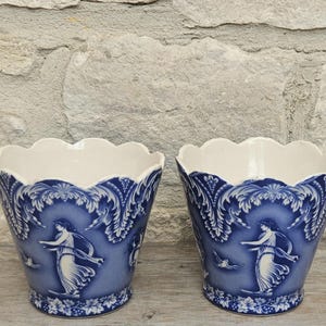 May include: A pair of ceramic planters featuring a scalloped rim and a deep blue and white transferware pattern. The design depicts classical figures in flowing robes surrounded by ornate foliate motifs and decorative borders on the exterior.