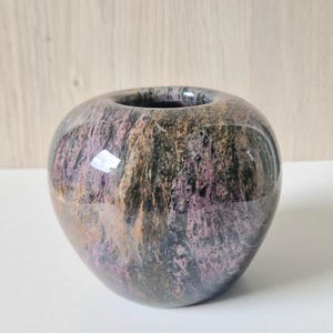 Puede incluir: Un pequeño jarrón redondo de piedra pulida con una abertura ancha. El jarrón tiene un patrón moteado de tonos morados, marrones y negros. La superficie es brillante, reflejando la luz. El jarrón probablemente se utiliza con fines decorativos.