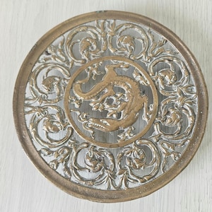 Puede incluir: Salvamanteles redondo de metal ornamentado con un diseño detallado. El salvamanteles presenta un círculo central con una figura de dragón, rodeado por un intrincado patrón floral y de volutas. El metal tiene un acabado dorado y plateado.