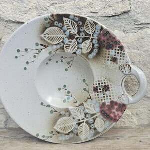 May include: A round ceramic serving plate with a single side handle, featuring a speckled off-white glaze. The surface is decorated with painted botanical motifs of leaves and berries in shades of blue, green, and reddish-brown, alongside a grid pattern.