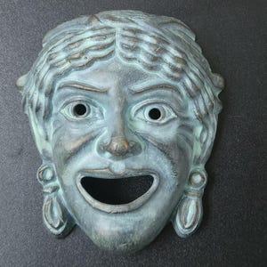 May include: A bronze-colored theatrical mask with a smiling expression. The mask has open eye sockets and a wide, open mouth. The hair is styled in waves, and the mask has decorative elements on the sides.