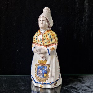 May include: A ceramic figurine of a woman wearing a white head covering, a yellow and red patterned shawl, and a white dress with a blue and gold coat of arms. The figurine is standing on a black surface.
