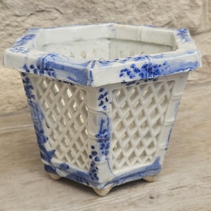 May include: A white ceramic planter with a hexagonal shape and an open lattice design. The planter features blue painted floral and bamboo-like patterns along the edges and corners, with small feet at the base for support and stability.