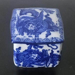 May include: A small, rectangular, lidded box with a blue and white design. The box features a detailed illustration of a mythical creature on both the lid and the base. The design is reminiscent of traditional Asian porcelain.