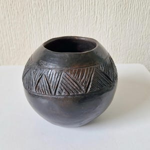 Puede incluir: Una pequeña olla de barro redonda y negra con un patrón geométrico tallado en la superficie.