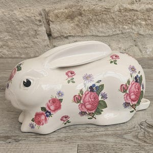 May include: A white ceramic bunny figurine with a glossy finish. The bunny is decorated with pink roses, blue and purple flowers, and green leaves. The bunny has black eyes and is sitting on a wooden surface.