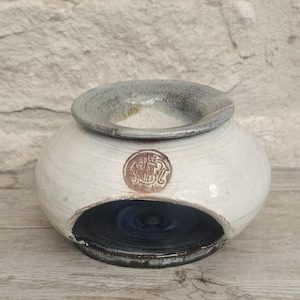 May include: A round, off-white ceramic oil burner with a wide, textured gray rim at the top. The body features a circular brown stamp with an embossed design. A large, arched opening at the base reveals a dark blue, glossy interior basin.