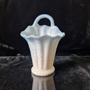 Puede incluir: Una cesta de cerámica blanca con un borde azul y un asa. La cesta tiene un patrón tejido y está diseñada para contener objetos pequeños.