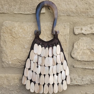 May include: A dark brown fabric handbag featuring a U-shaped handle with a marbled brown and black pattern. The exterior is covered in rows of overlapping, teardrop-shaped white shell pieces attached with dark brown thread and small dark beads.