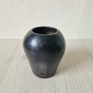 May include: A small, black vase with a rounded body and a wide opening. The vase has a smooth, matte finish and a slightly tapered base. It is a decorative item that can be used to hold flowers or other small objects.