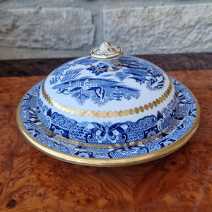 Puede incluir: Mantequillera de cerámica azul y blanca con tapa, con un diseño detallado de paisaje. El plato tiene un borde y detalles dorados, añadiendo un toque de elegancia. La tapa tiene un pequeño asa decorativa.