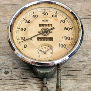 May include: A vintage British Jaeger circular speedometer gauge with a chrome bezel and a cream-coloured face. The dial features black numerals from 0 to 100, a central odometer display, and a smaller inset clock dial at the bottom with two metal needles.
