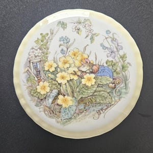 May include: A round decorative plate with a yellow border. The center features a detailed illustration of a mouse in a blue coat, nestled among yellow primroses and other flowers. The plate is signed "Jill Barklem".