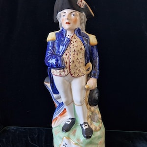 Puede incluir: Figura de cerámica de un hombre con abrigo azul, calzones blancos y sombrero negro. La figura está sobre una base verde con el nombre "Nelson" escrito. El abrigo tiene charreteras doradas y el sombrero un escudo.