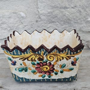 May include: A rectangular ceramic planter with a jagged, zig-zag top edge painted in dark brown. The exterior features a cream-coloured base with hand-painted floral motifs in yellow, teal, and maroon, accented by dark blue vertical stripes.