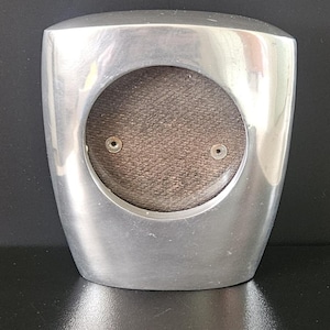 May include: A brushed silver photo frame with a rounded rectangular shape. The frame has a circular opening with a brown fabric backing and two small silver rivets. The frame is designed to stand upright.