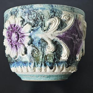 Puede incluir: Una maceta de cerámica con un diseño texturizado y multicolor. La maceta presenta un gran motivo floral morado y blanco y patrones decorativos de hojas arremolinadas. El fondo tiene tonos de azul, verde y negro.