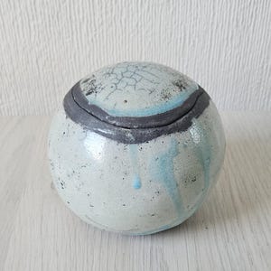 Peut inclure: Un pot en céramique blanche avec un couvercle, recouvert d'un glaçage bleu et gris. Le pot a une finition craquelée et une forme arrondie.