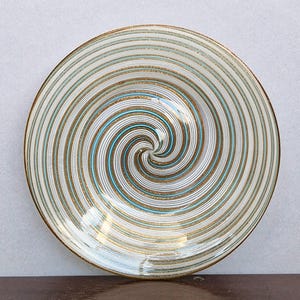Könnte beinhalten: Ein runder Keramikteller mit einem wirbelnden Spiralmuster, das von der Mitte ausstrahlt. Das Design besteht aus abwechselnden dünnen Linien in Metallic-Gold, hellem Türkis und sanftem Weiß, die eine kontinuierliche kreisförmige Bewegung erzeugen.