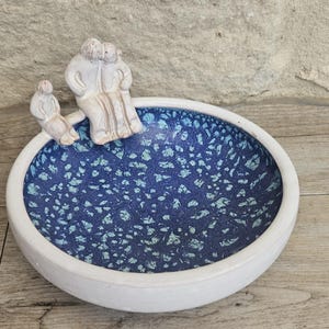 Peut inclure: Un bol en céramique avec un extérieur blanc et un intérieur bleu moucheté. Le bol présente un élément décoratif de trois personnages. Les personnages sont de couleur claire et semblent s'embrasser. Le bol est posé sur une surface en bois.