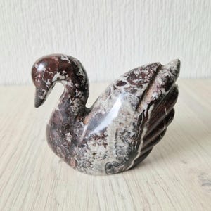 Könnte beinhalten: Eine geschnitzte Schwan-Skulptur aus Stein. Der Schwan ist hauptsächlich dunkelrotbraun mit weißen und grauen Marmorierungen. Die Skulptur hat eine glatte, polierte Oberfläche und steht auf einer hellen Holzoberfläche.