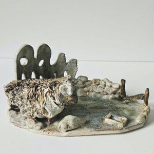 May include: A ceramic sculpture of a sheep in a pen. The sheep is off-white with textured fur. The pen has a stone wall and a small trough. The sculpture is on a white surface.